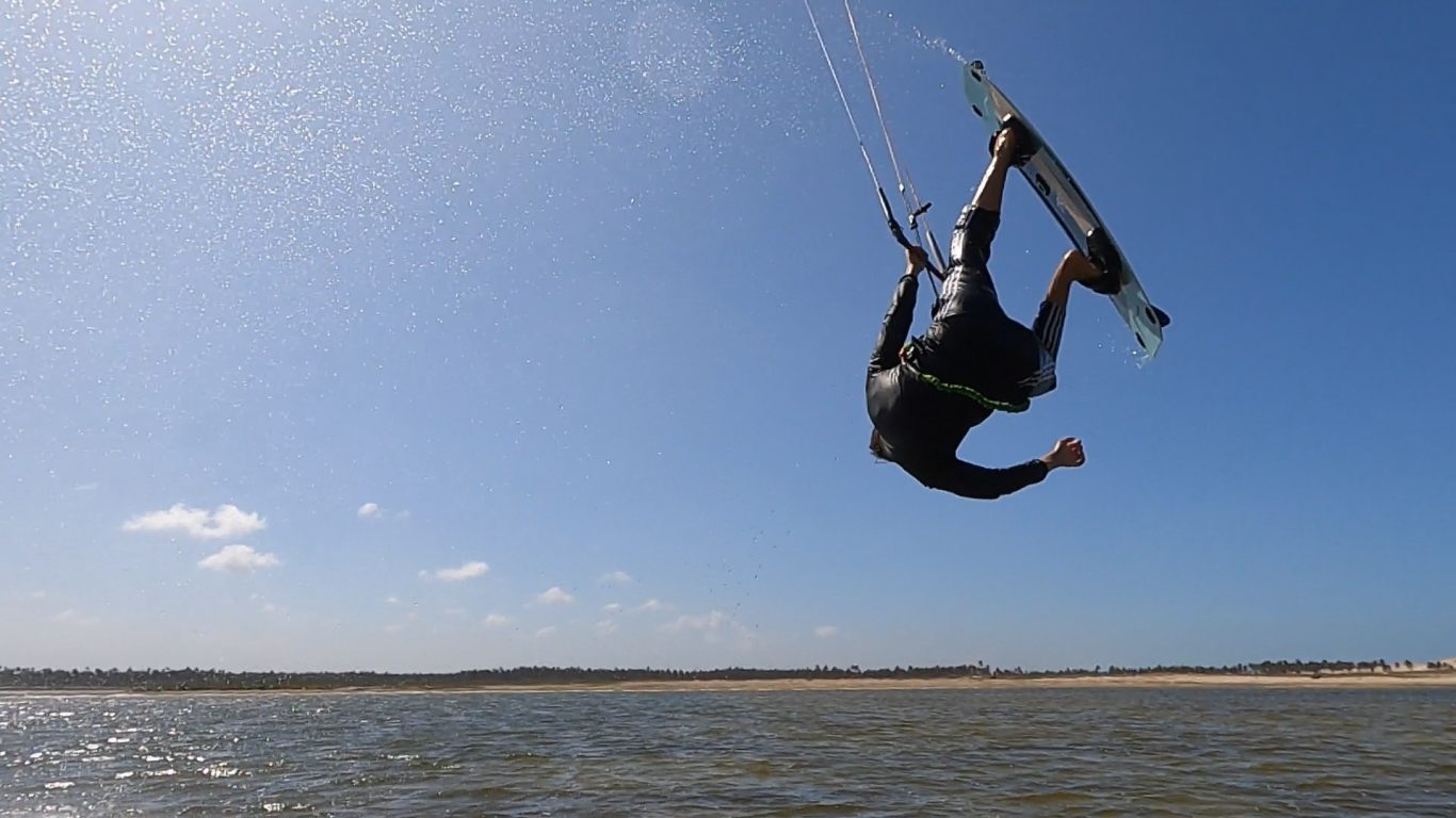 Kitesurfer springt über das Wasser, blauer Himmel und flaches Ufer im Hintergrund.