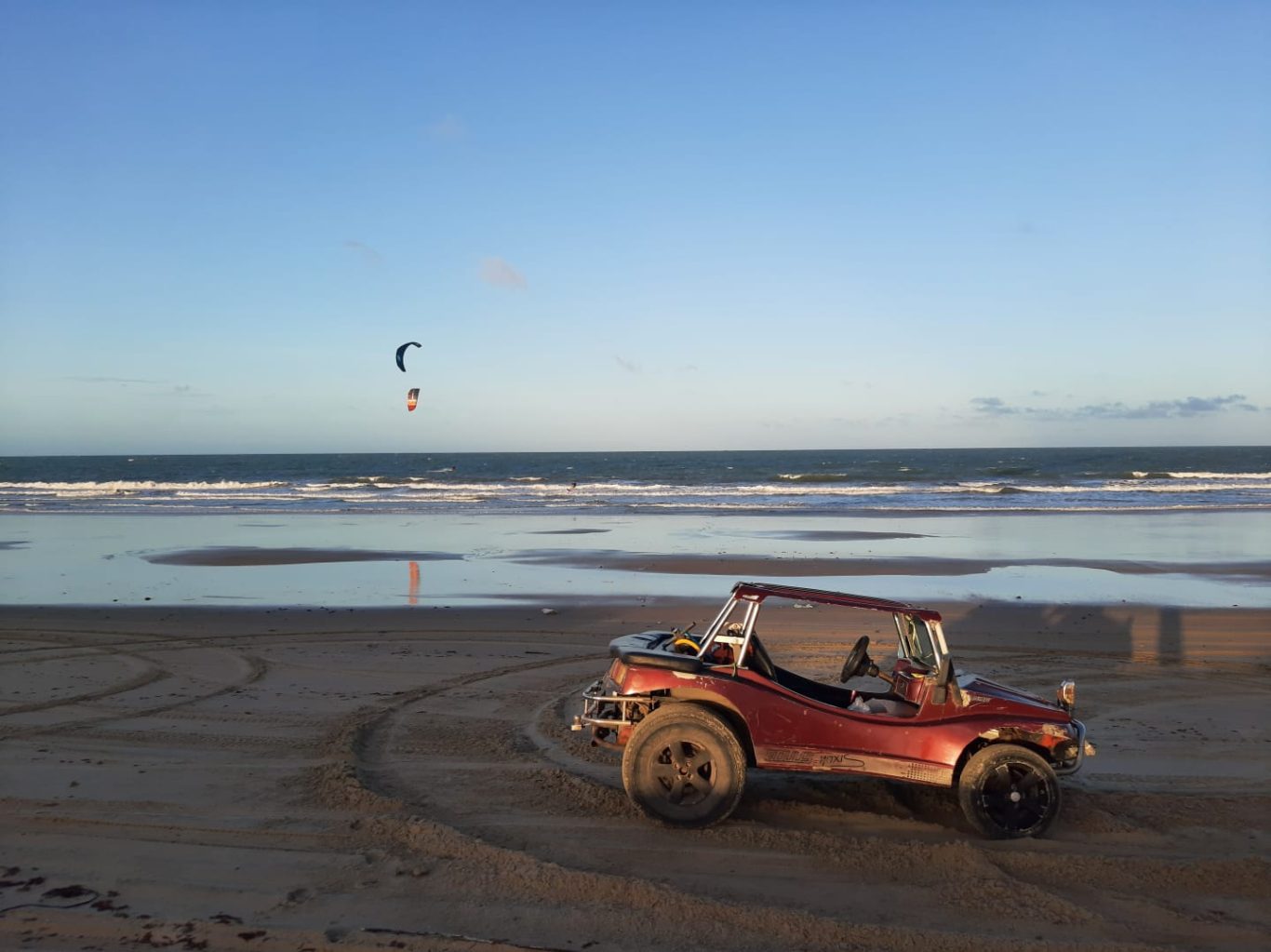 Roter Buggy am Strand mit Kitesurfern im Hintergrund und Wellen.