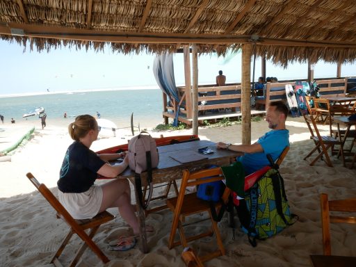 Zwei Personen sitzen am Strand in einem Schattenbereich mit Blick auf das Wasser.