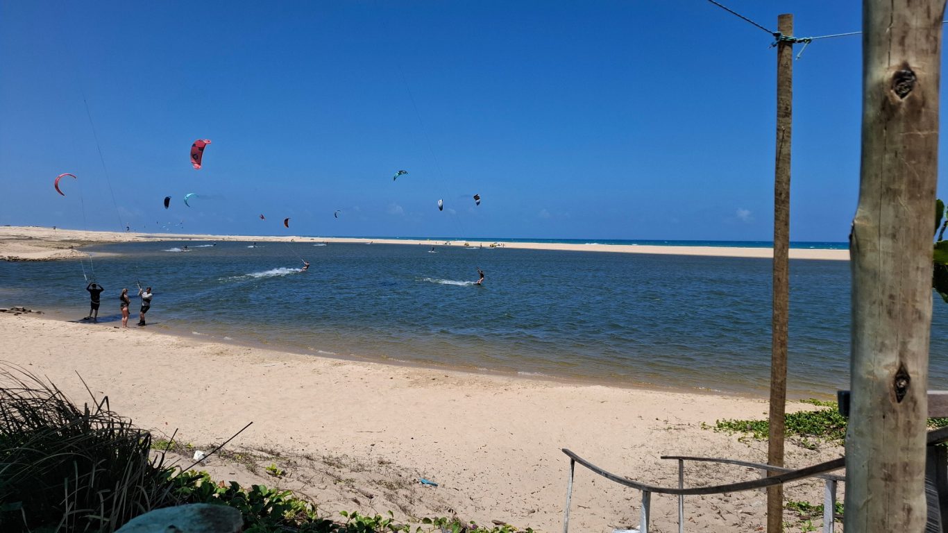 Strand mit flachem Wasser, Kitesurfer in der Ferne und blauer Himmel.