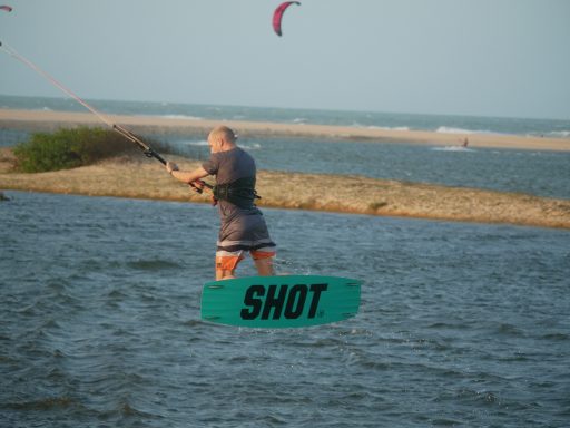 Ein Mann beim Kitesurfen springt übers Wasser, auf der Rückseite vom Board steht 'SHOT'.