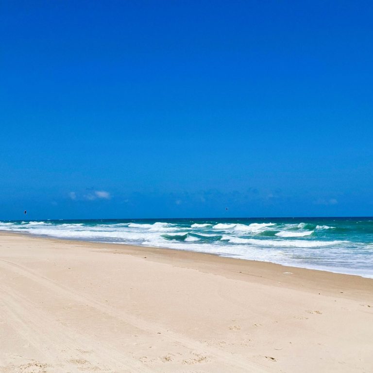 Weißer Sandstrand mit Wellen und klarem, blauem Himmel.