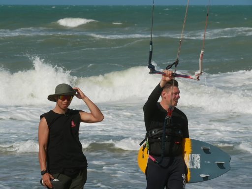 Zwei Surfer am Strand, einer hält ein Kitesurf-Brett und lächelt. Wellen im Hintergrund.