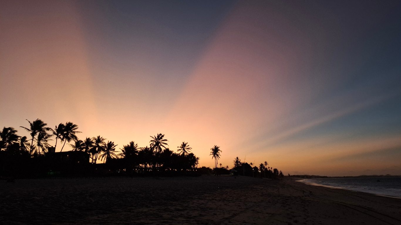 Sonnenuntergang am Strand mit Palmen und sanften Lichtstrahlen am Himmel.