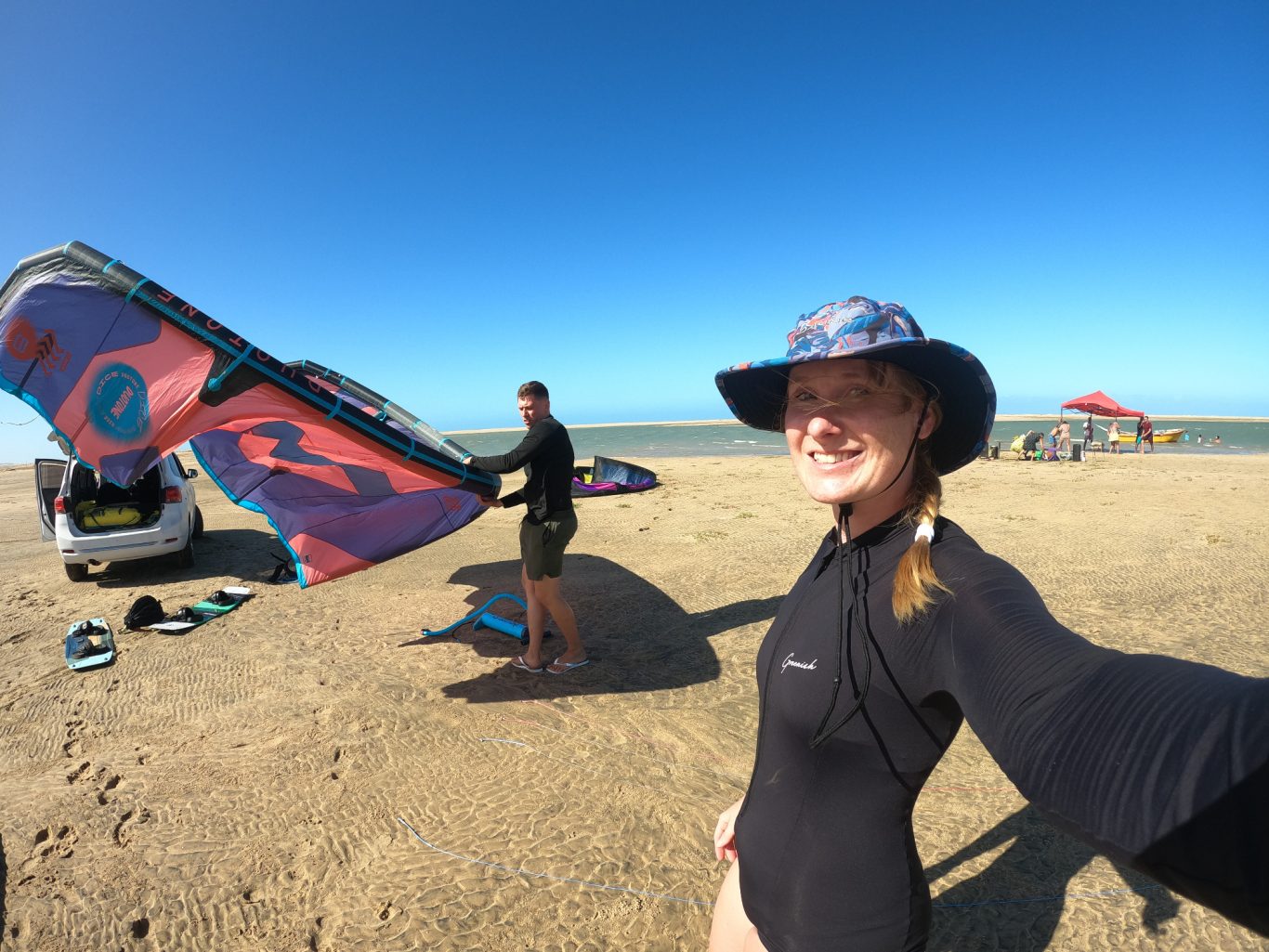 Zwei Personen am Strand, eine mit Hut und die andere mit einem Kite. Sonniger Himmel.