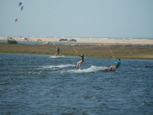 Drei Personen beim Kitesurfen auf einem Gewässer, mit Strand im Hintergrund.