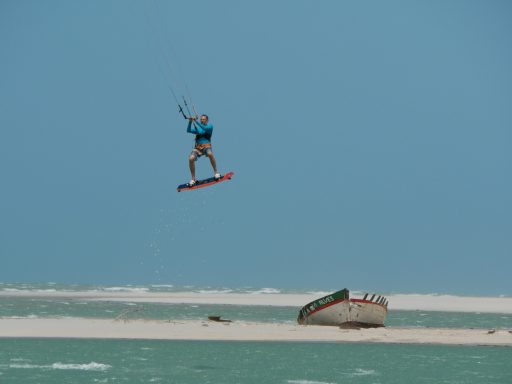 Kitesurfer springt über das Wasser vor einer Küste mit einem Boot im Hintergrund.