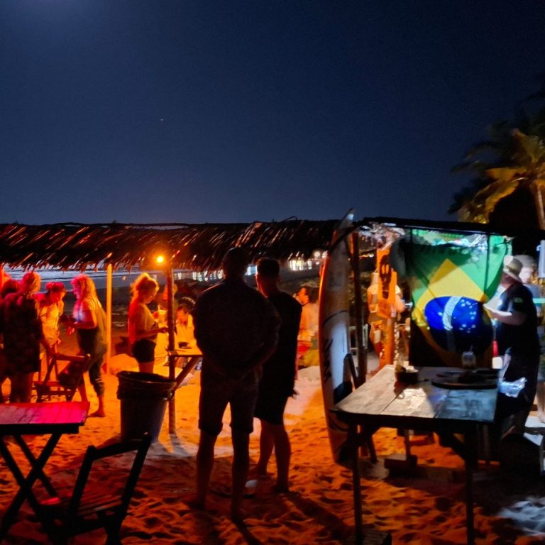Menschen am Strand bei Nacht an einer Bar, umgeben von Lichtern und Palmen, entspannte Atmosphäre.