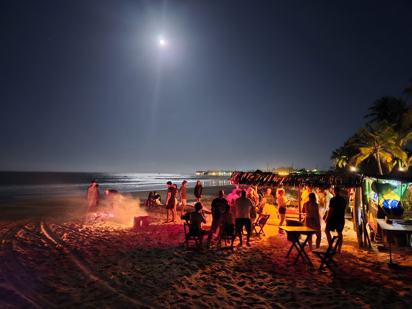 Nachtstrand mit Menschen, Feuerstellen und Mondlicht über dem Meer.