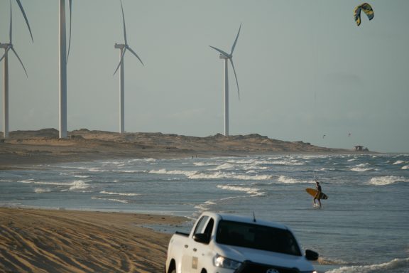 Ein Surfer steht am Strand, während Windräder im Hintergrund und ein Auto im Vordergrund zu sehen sind.