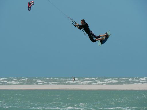 Kitesurfer springt über das Wasser, mit weitem Strand im Hintergrund.